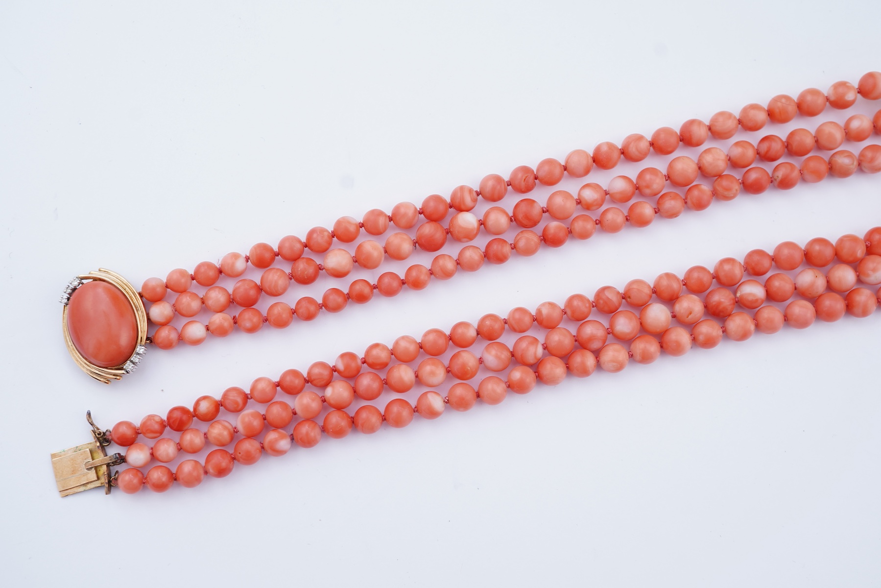 A coral necklace, composed of three strands of coral beads measuring approximately 5.1-6.6mm diameter, to a gold clasp set with a coral cabochon and brilliant-cut diamonds, mounted in gold, length 45.5cm, stamped 14K, ca